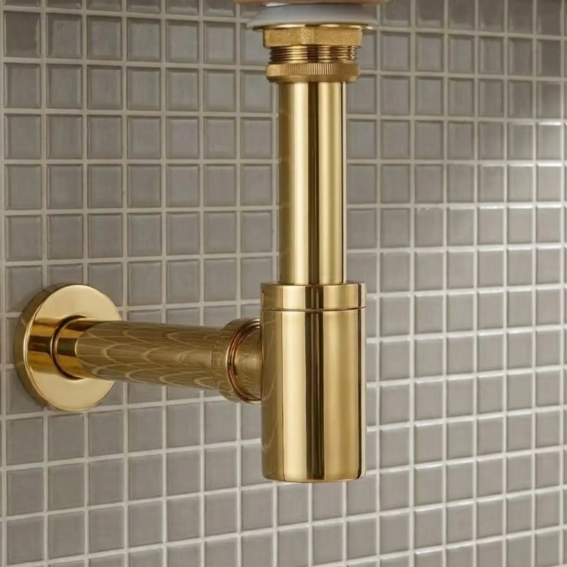 Front view of 1-1/4 inch unlacquered brass round bottle P-trap with box flange mounted on gray mosaic tile bathroom wall
