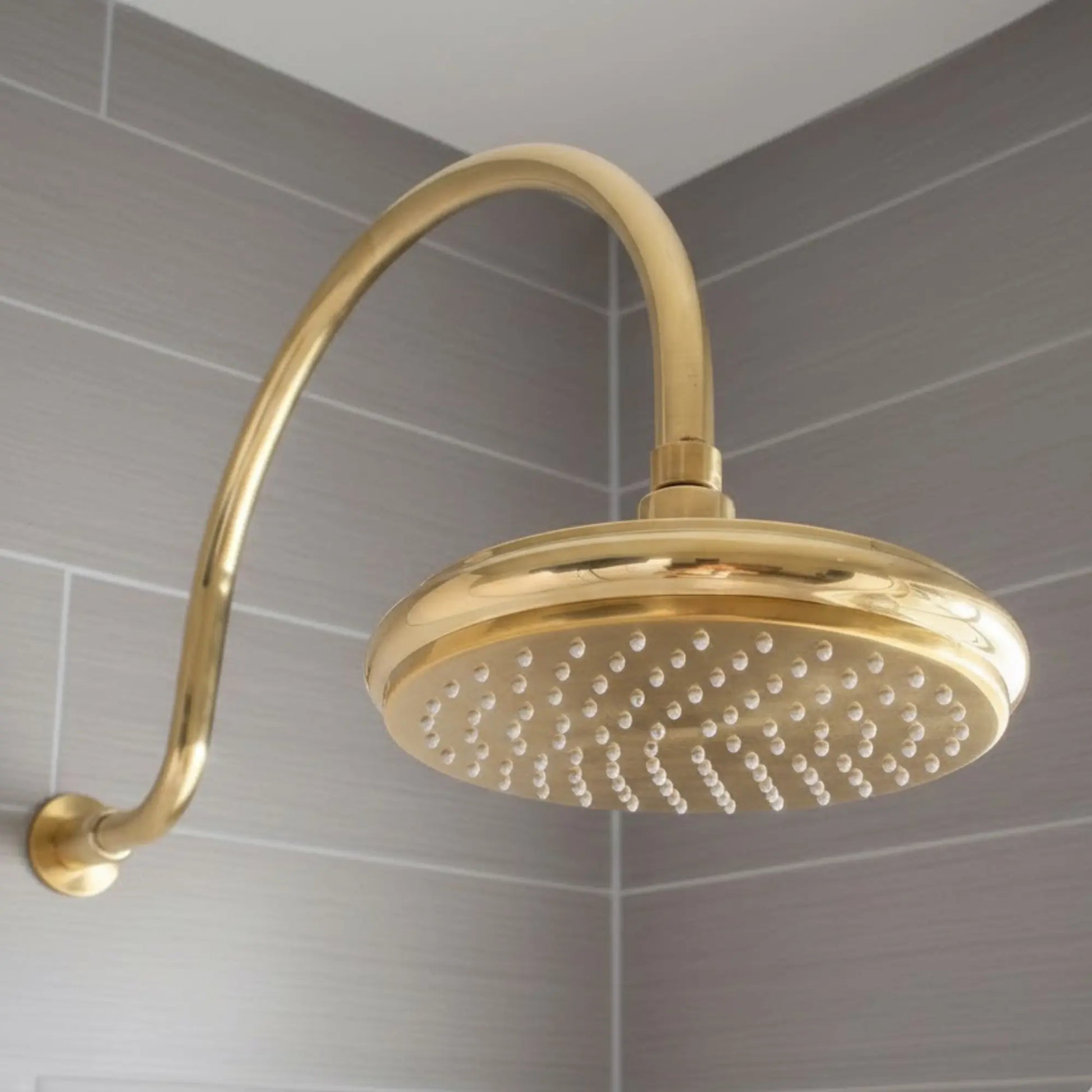 Close-up unlacquered brass rainfall shower head mounted on curved wall arm in modern tiled bathroom