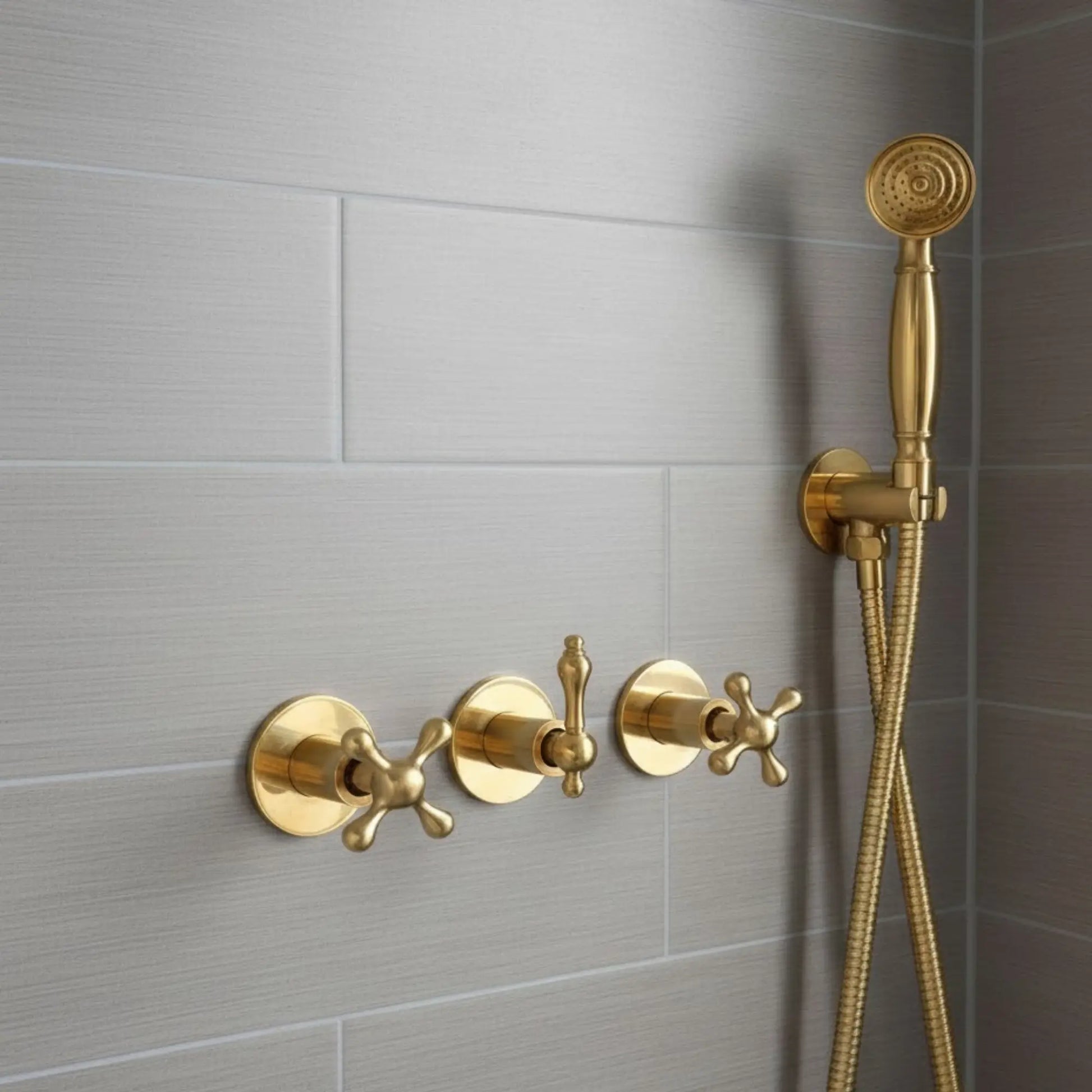 Close-up of unlacquered brass shower mixer with cross handle controls and handheld shower sprayer installed on gray tiled bathroom wall