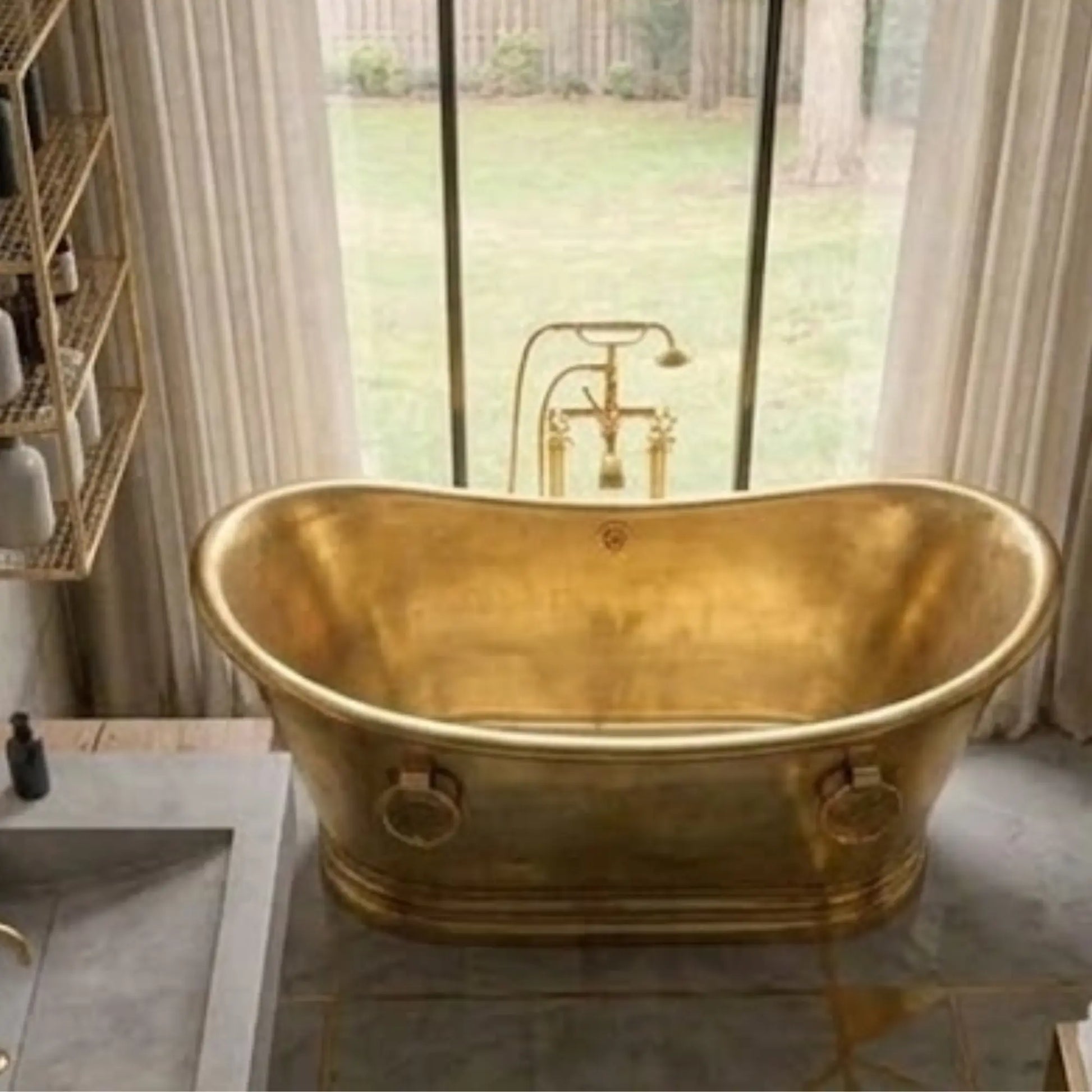 A freestanding oval bathtub in a warm brushed gold finish, positioned in a bathroom with a large window, sheer curtains, and a matching gold faucet.