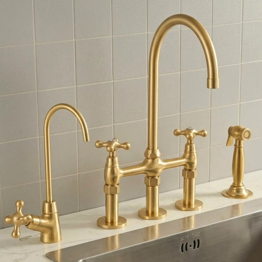 A brushed gold bridge faucet set with a high-arc main spout, a secondary side spout with a cross handle, a separate hot/cold valve, and a handheld sprayer, installed on a white marble countertop against a light gray tiled backsplash.