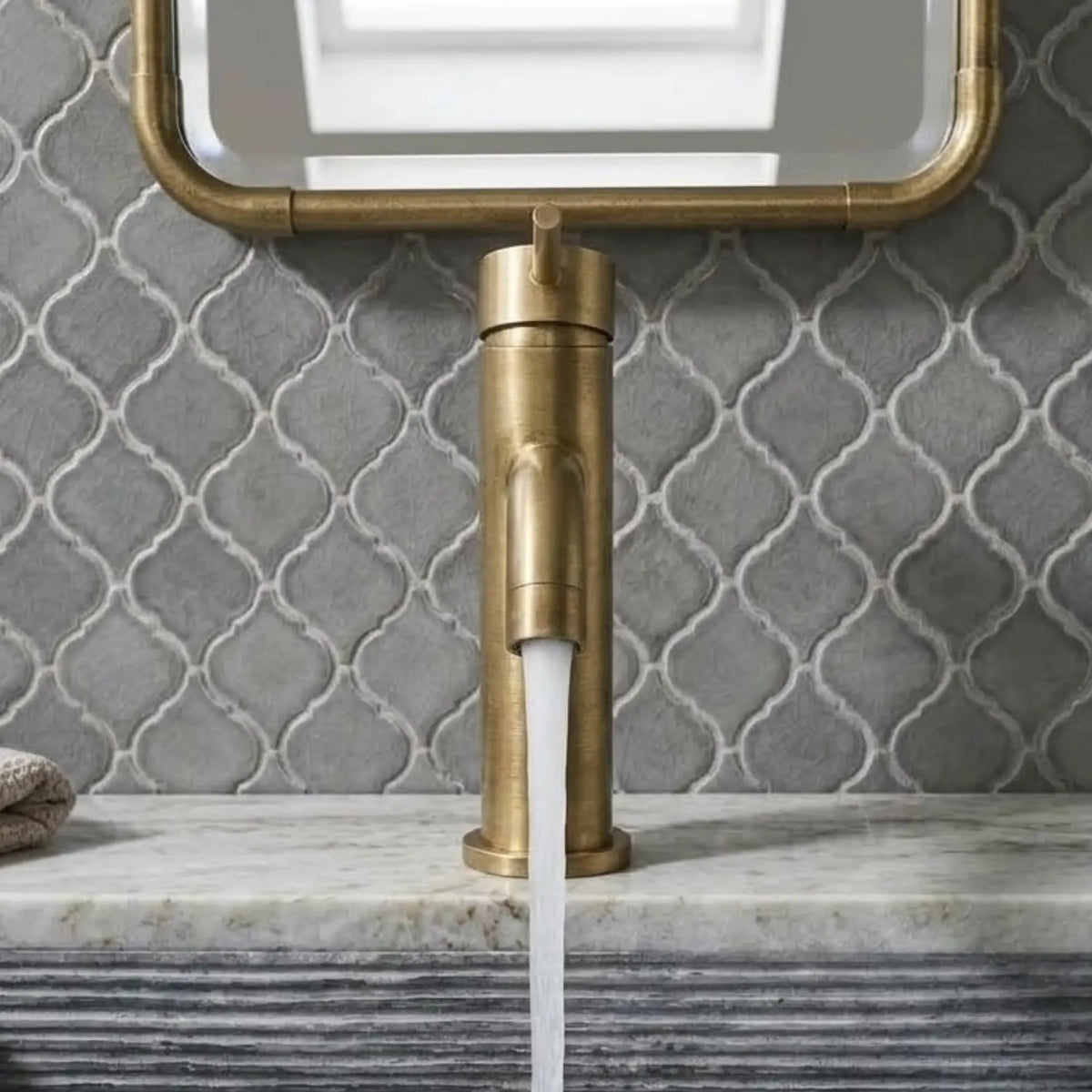 Front view of an unlacquered brass single-hole faucet with water flowing into a fluted gray sink, set against a gray arabesque tile wall and matching brass-framed mirror.