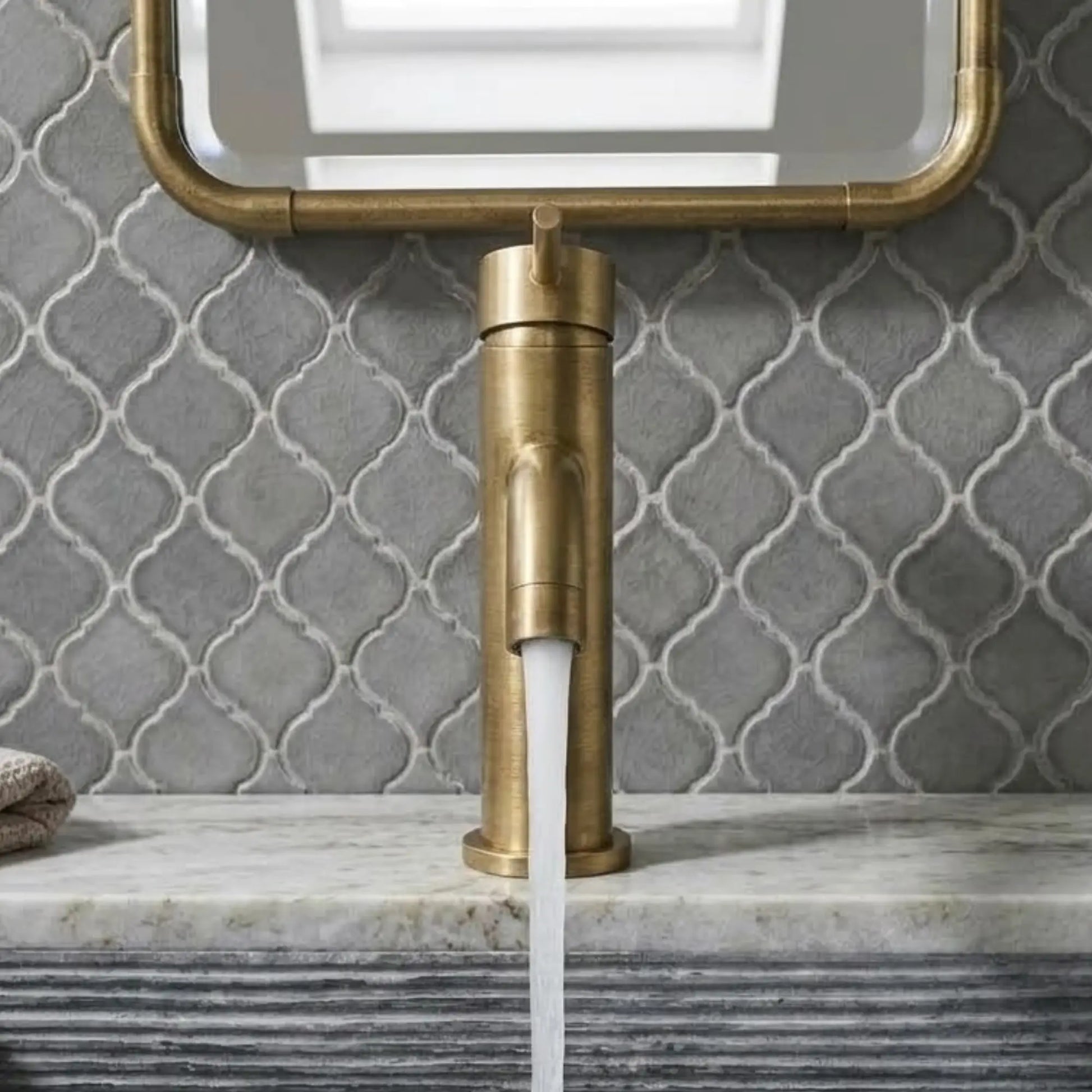 Front view of an unlacquered brass single-hole faucet with water flowing into a fluted gray sink, set against a gray arabesque tile wall and matching brass-framed mirror.