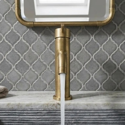 Front view of an unlacquered brass single-hole faucet with water flowing into a fluted gray sink, set against a gray arabesque tile wall and matching brass-framed mirror.