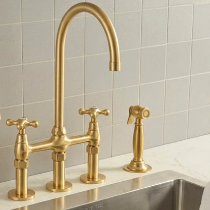 A three-hole bridge faucet set in brushed gold brass, featuring a high-arc main spout, two cross-handle hot and cold valves, and a matching side spray nozzle, installed on a white marble countertop against a light gray tiled backsplash.