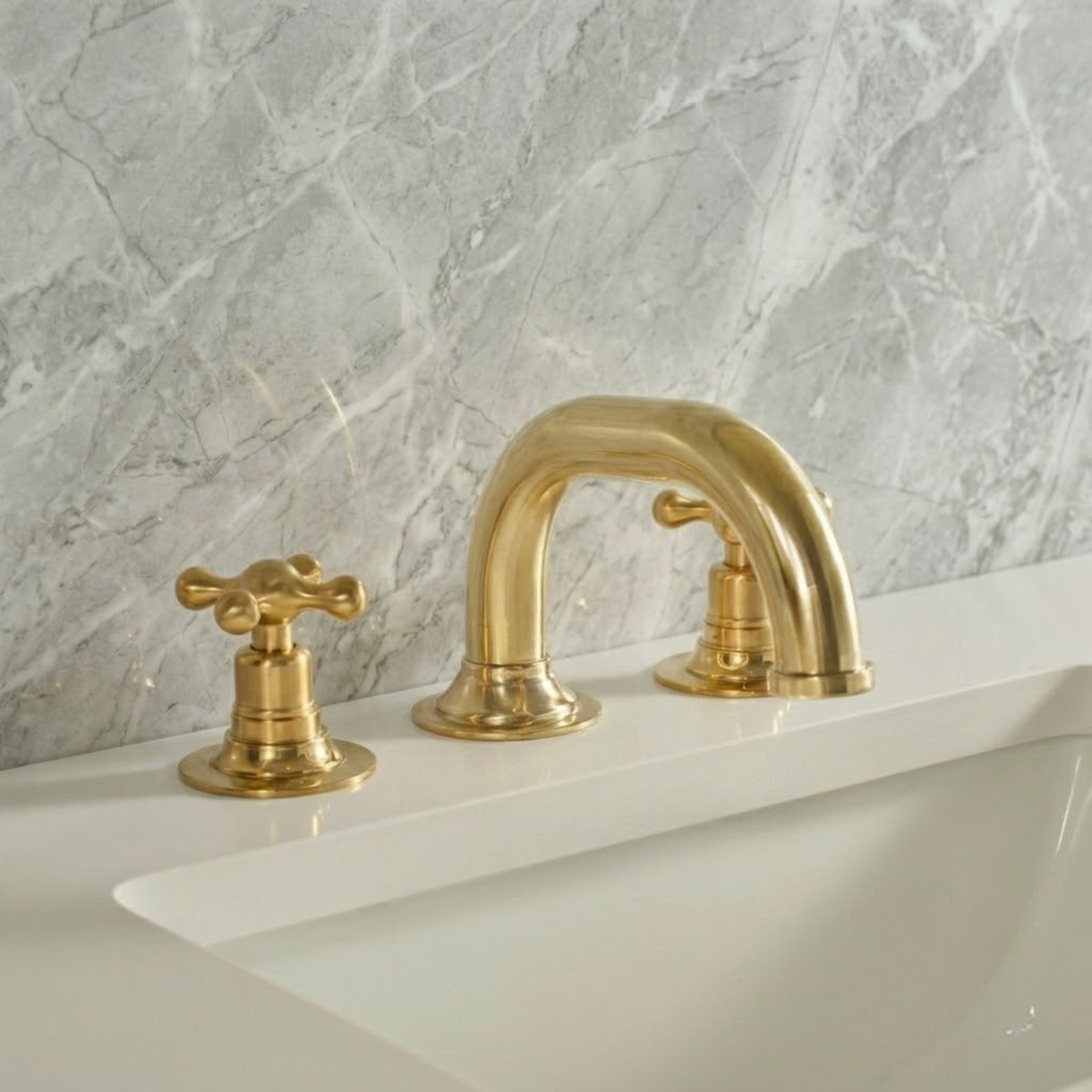 Luxury unlacquered brass 3-hole widespread bathroom faucet with cross handles mounted on a white vanity sink with light gray marble wall