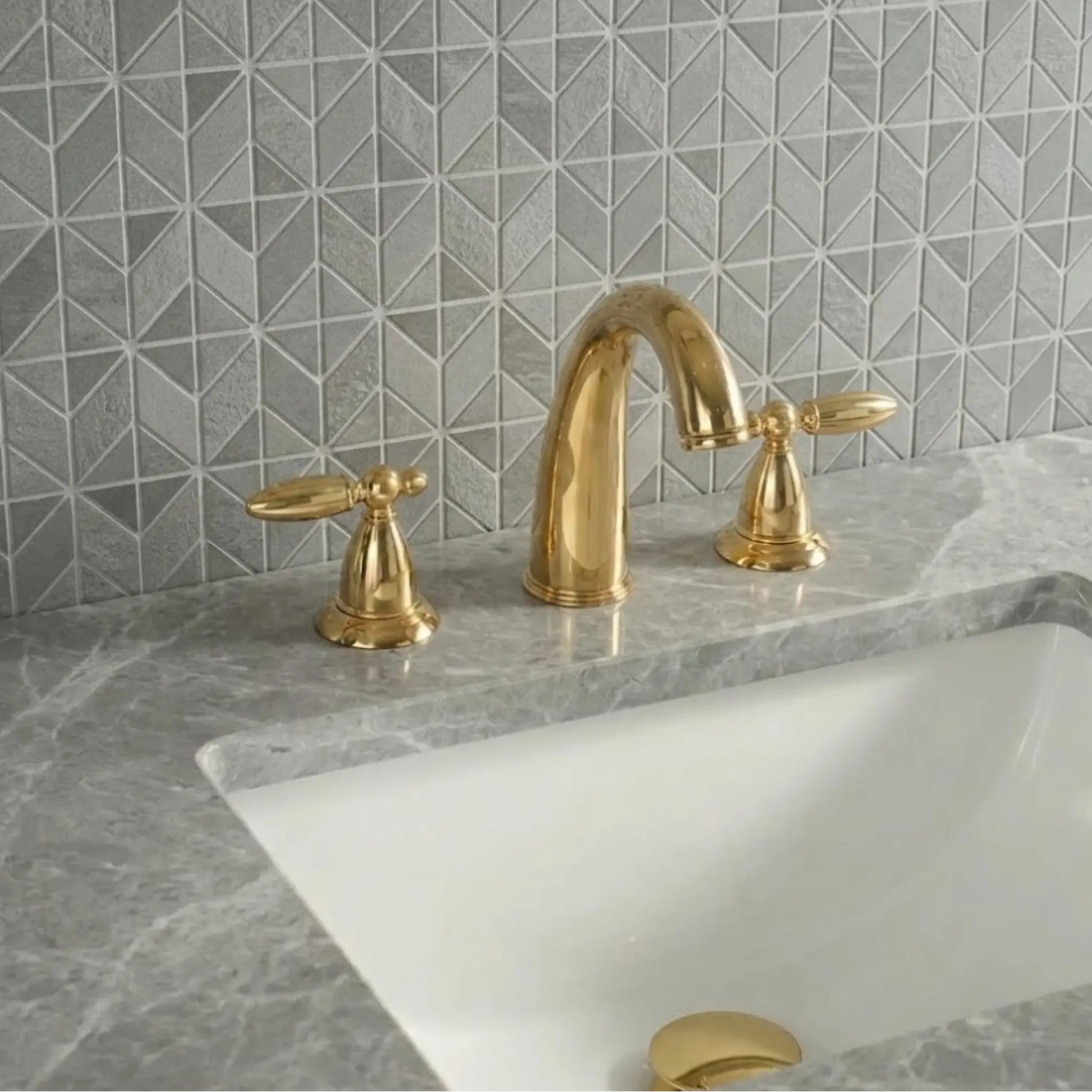 Sella unlacquered brass widespread faucet close-up showing high-arc gooseneck spout and cross handles on gray marble countertop with geometric tile backsplash