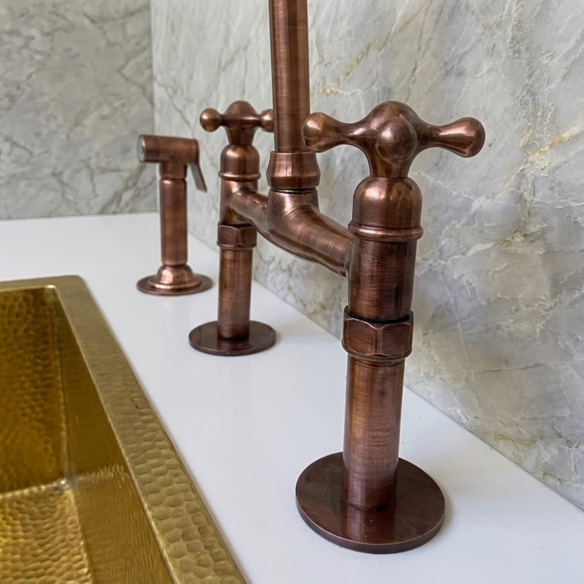 Close-up of solid copper bridge kitchen faucet cross handles and side sprayer next to brass farmhouse sink