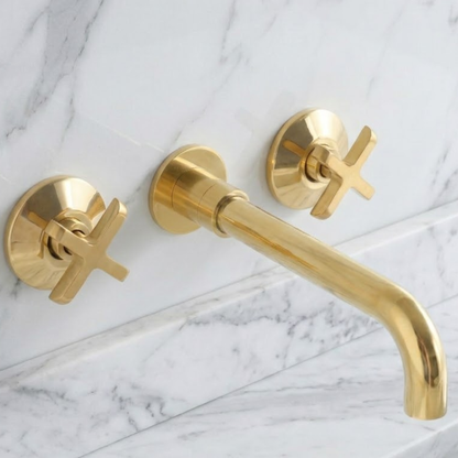 Solid unlacquered brass wall-mounted bathroom faucet with cross handles installed on a white marble backsplash above a brass sink, featuring a natural living finish.