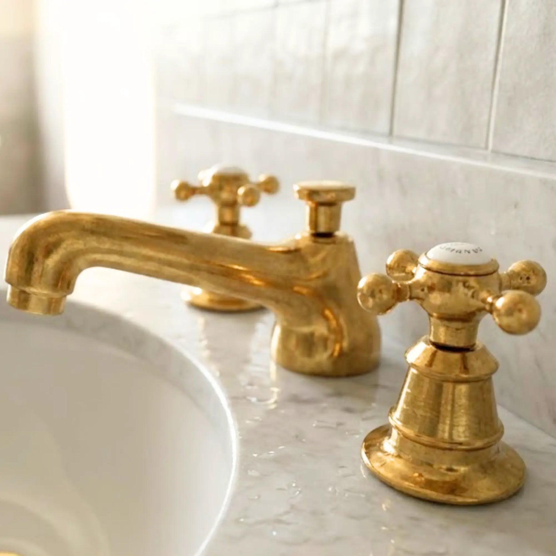 Unlacquered brass widespread bathroom faucet with cross handles on marble sink and light gray tiled wall