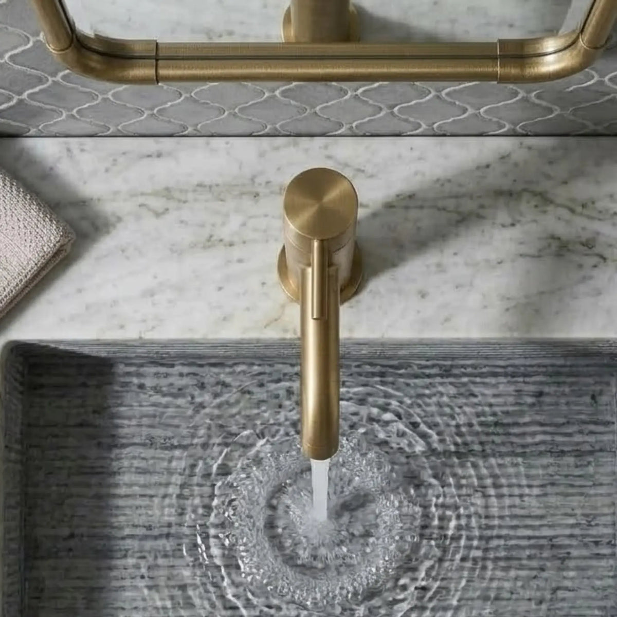 Overhead view of a modern unlacquered brass faucet pouring water into a gray linear-textured stone sink, showcasing a white marble countertop and brass mirror detail.
