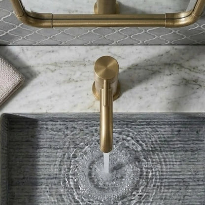 Overhead view of a modern unlacquered brass faucet pouring water into a gray linear-textured stone sink, showcasing a white marble countertop and brass mirror detail.