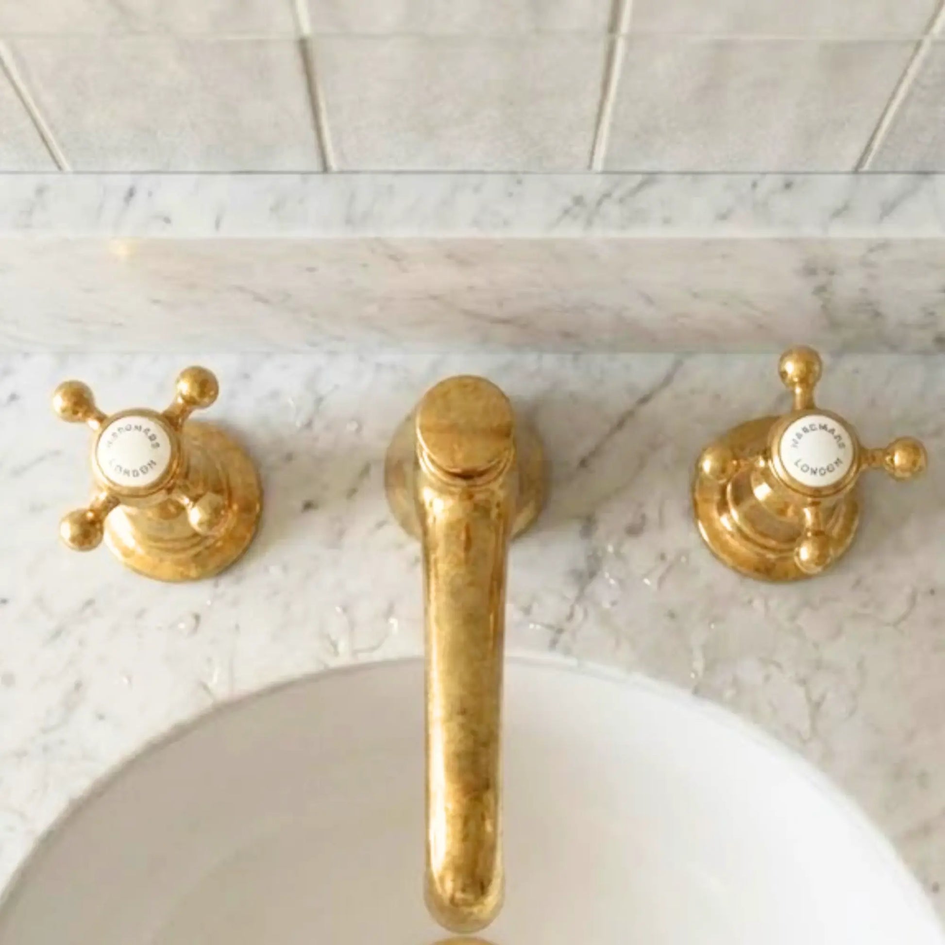Top view unlacquered brass widespread bathroom faucet with cross handles on white marble vanity and ceramic tile wall