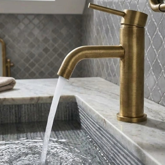 Modern unlacquered brass single-lever faucet with water flowing into a gray fluted stone sink against a grey patterned tile wall.