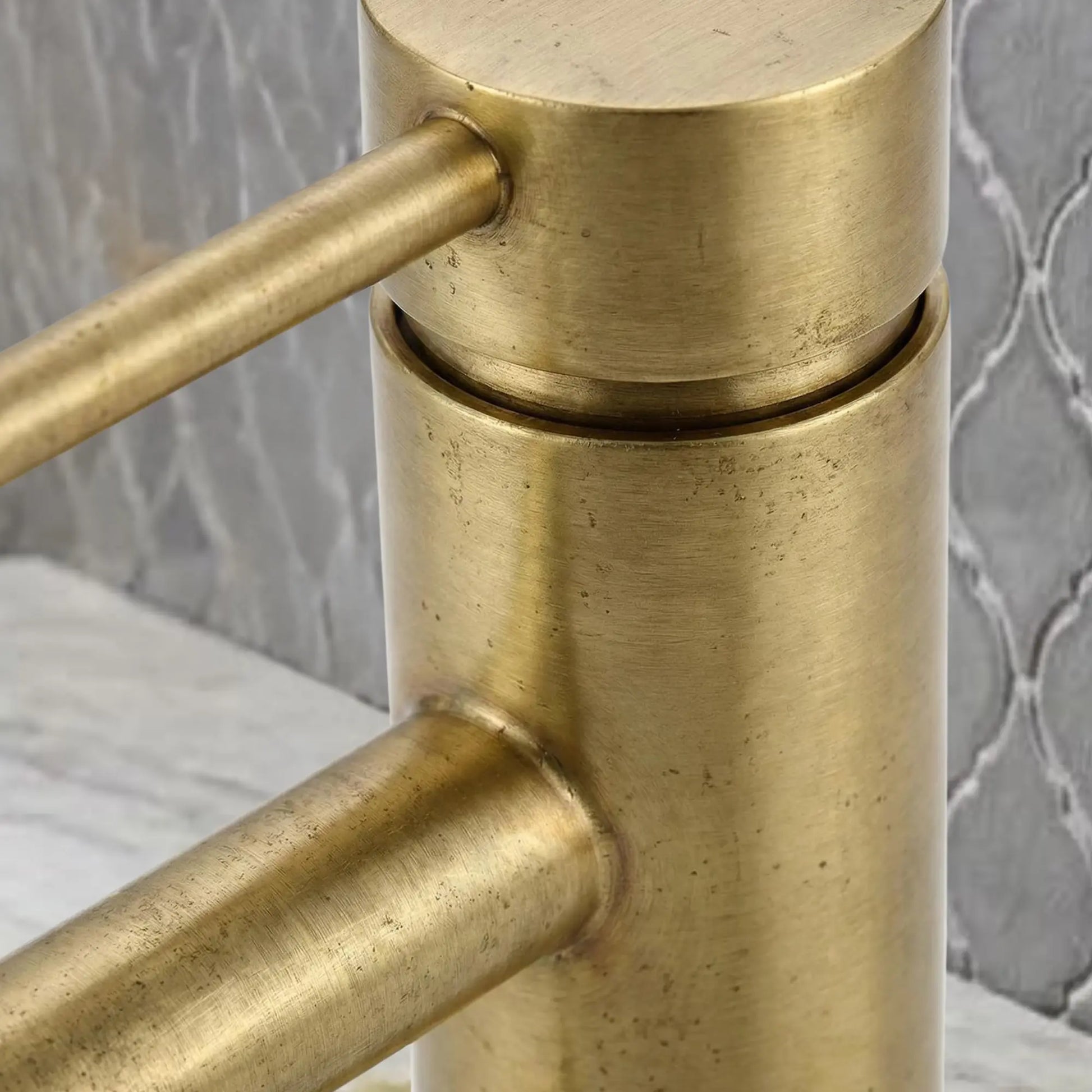 Extreme close-up of unlacquered brass faucet showing the authentic brushed metal texture and natural patina living finish against a blurred gray tile background.