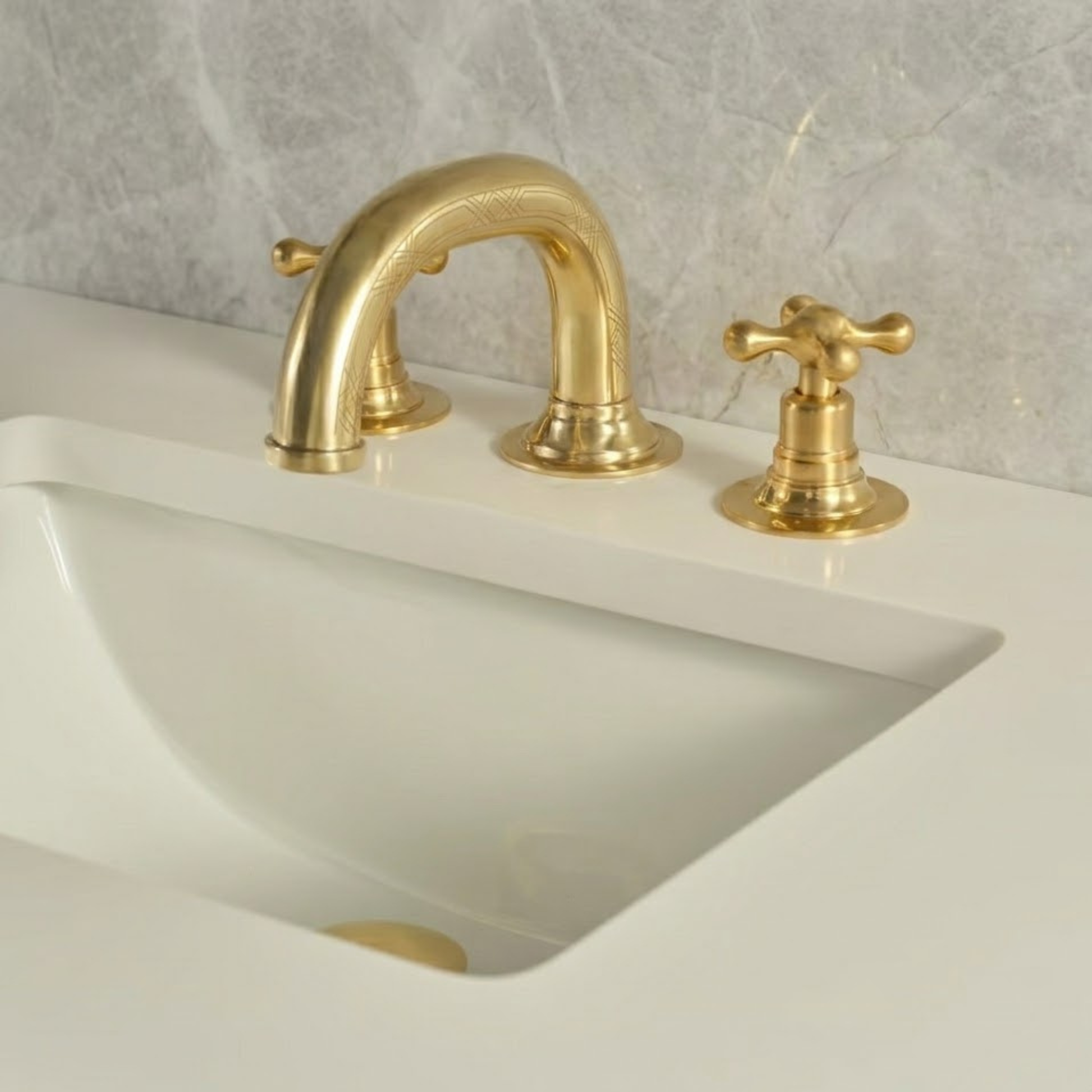 Solid unlacquered brass widespread bathroom faucet with cross handles and decorative engraved spout installed on a white sink with marble wall background