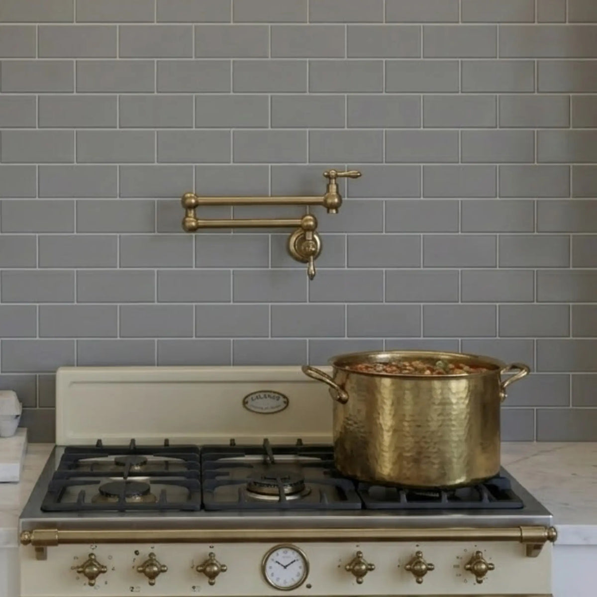 A wall-mounted unlacquered brass pot filler installed on a gray subway tile backsplash above a vintage-style cream gas stove with a large brass stockpot.