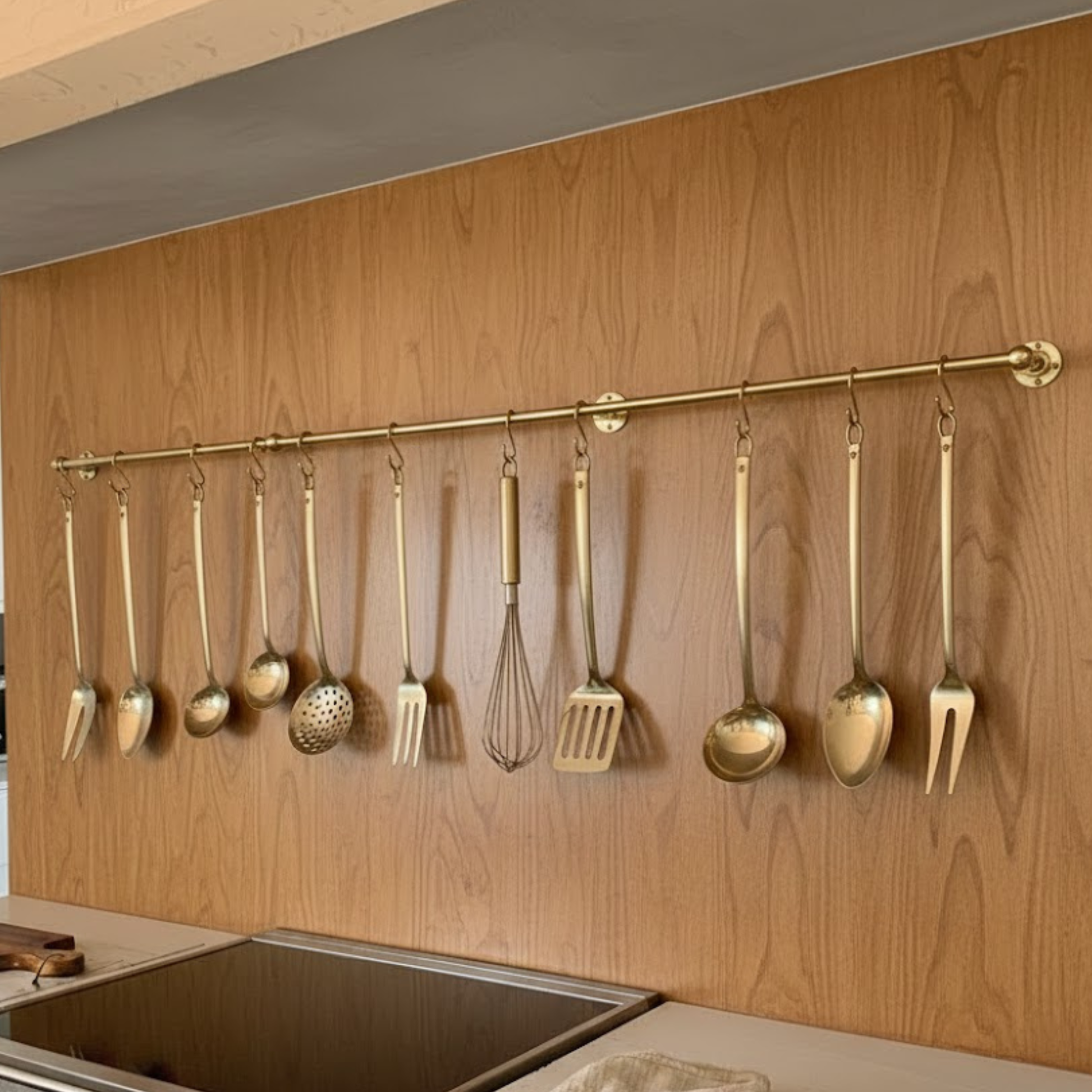 Unlacquered brass wall mounted pot rail with hooks holding kitchen utensils above sink