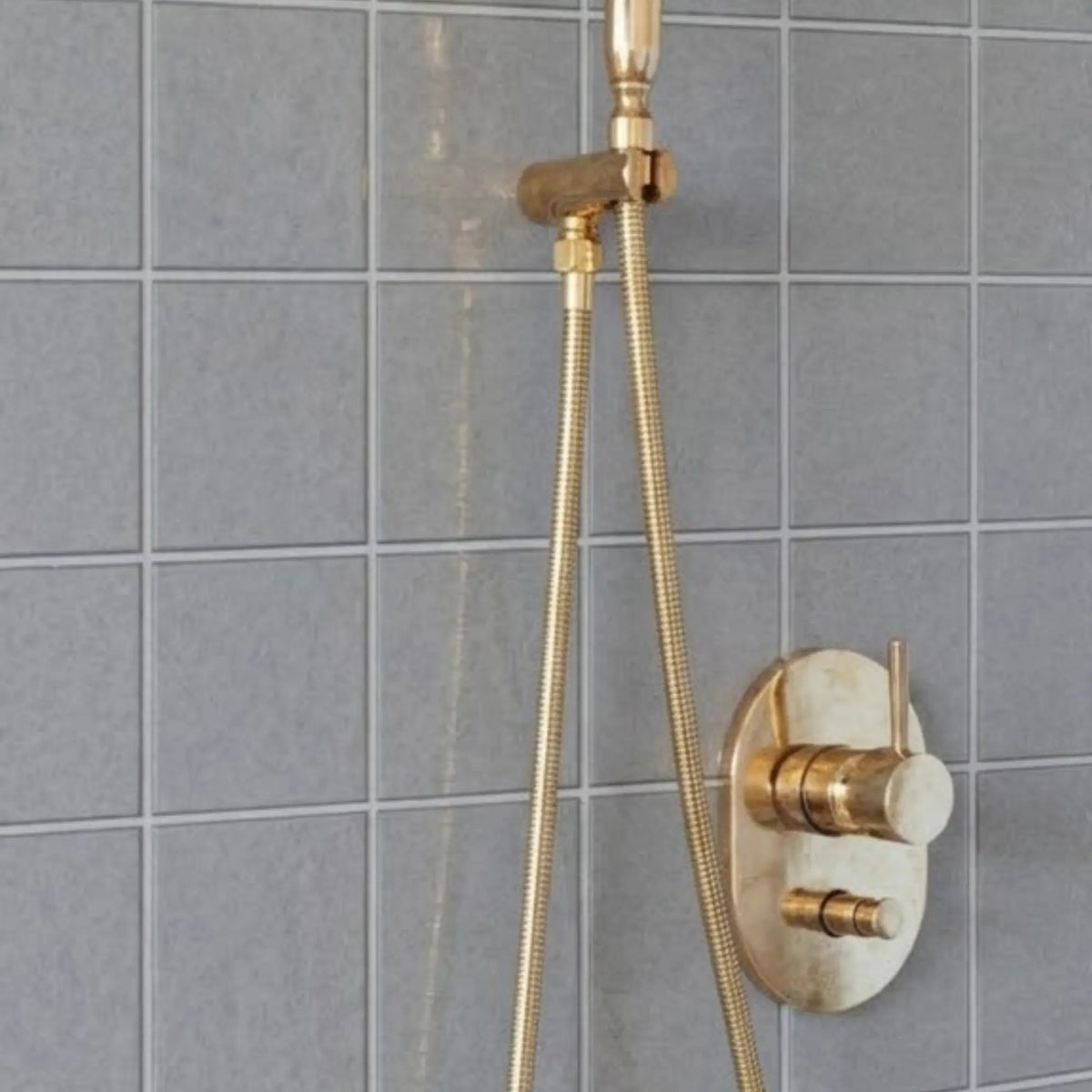 Close-up of unlacquered brass shower valve control and handheld shower hose installed on gray tiled bathroom wall