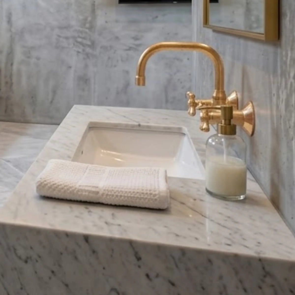 Unlacquered brass wall-mount faucet over white undermount sink on Carrara marble countertop with hand towel and soap dispenser