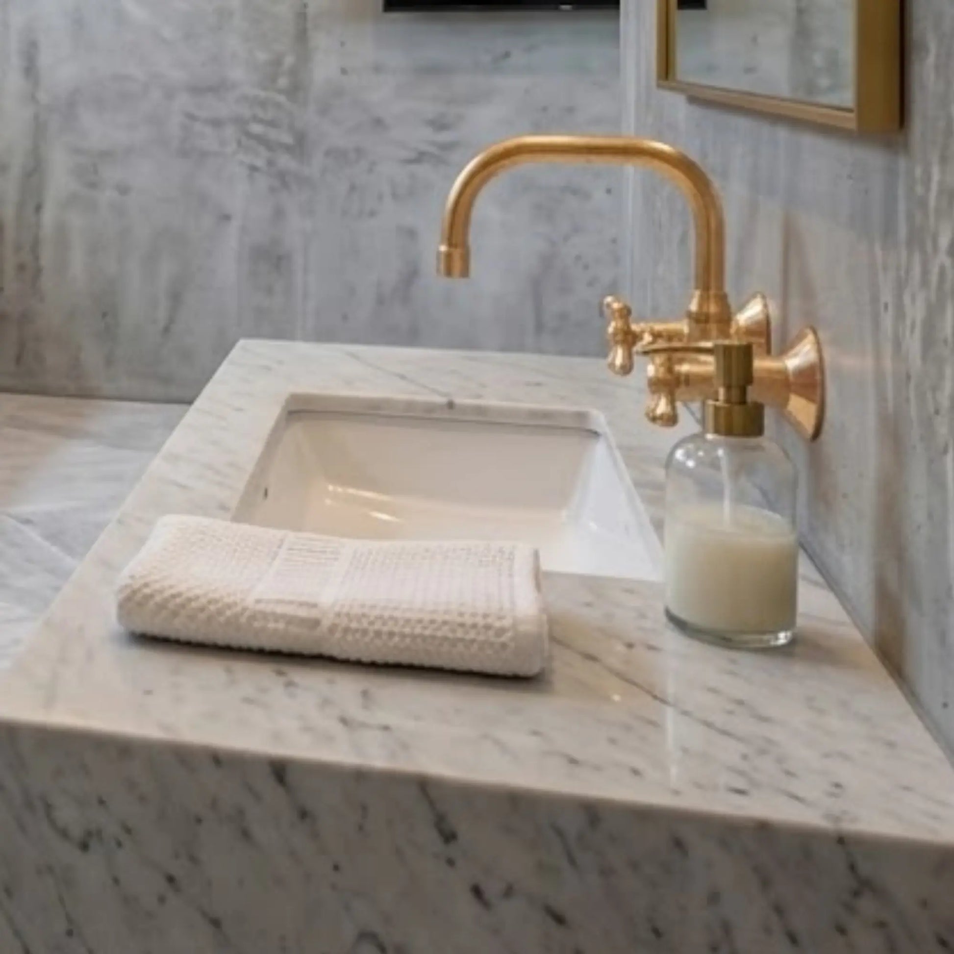Unlacquered brass wall-mount faucet over white undermount sink on Carrara marble countertop with hand towel and soap dispenser
