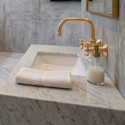 Unlacquered brass wall-mount faucet over white undermount sink on Carrara marble countertop with hand towel and soap dispenser