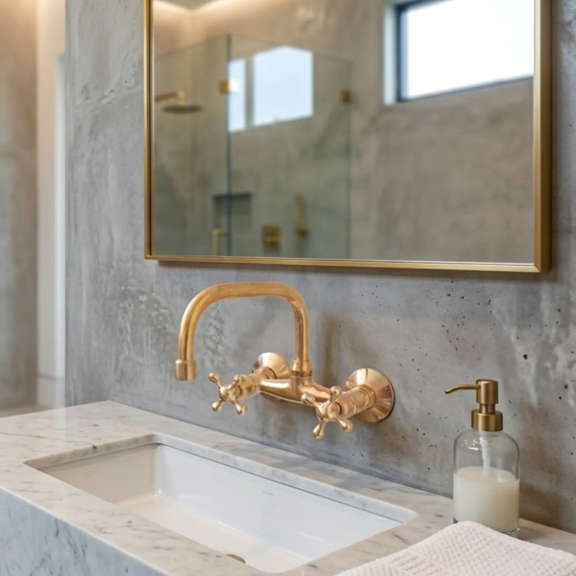 Unlacquered brass wall-mount faucet with cross handles on marble vanity, concrete wall, gold mirror and brass soap dispenser