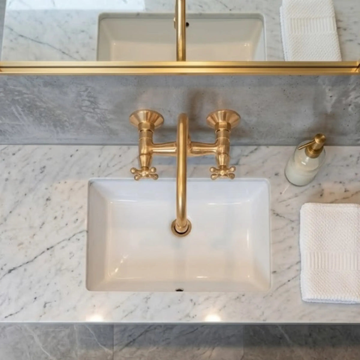 Unlacquered brass wall-mount faucet overhead view with cross handles above white undermount sink on Carrara marble vanity