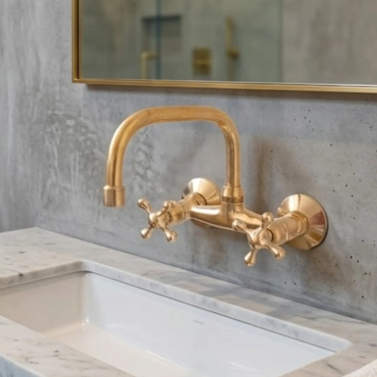 Unlacquered brass wall-mount kitchen faucet with cross handles above white undermount sink, raw concrete wall background