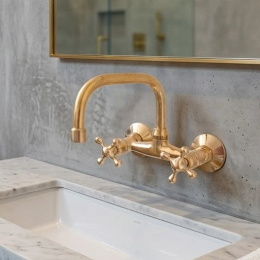 Unlacquered brass wall-mount kitchen faucet with cross handles above white undermount sink, raw concrete wall background