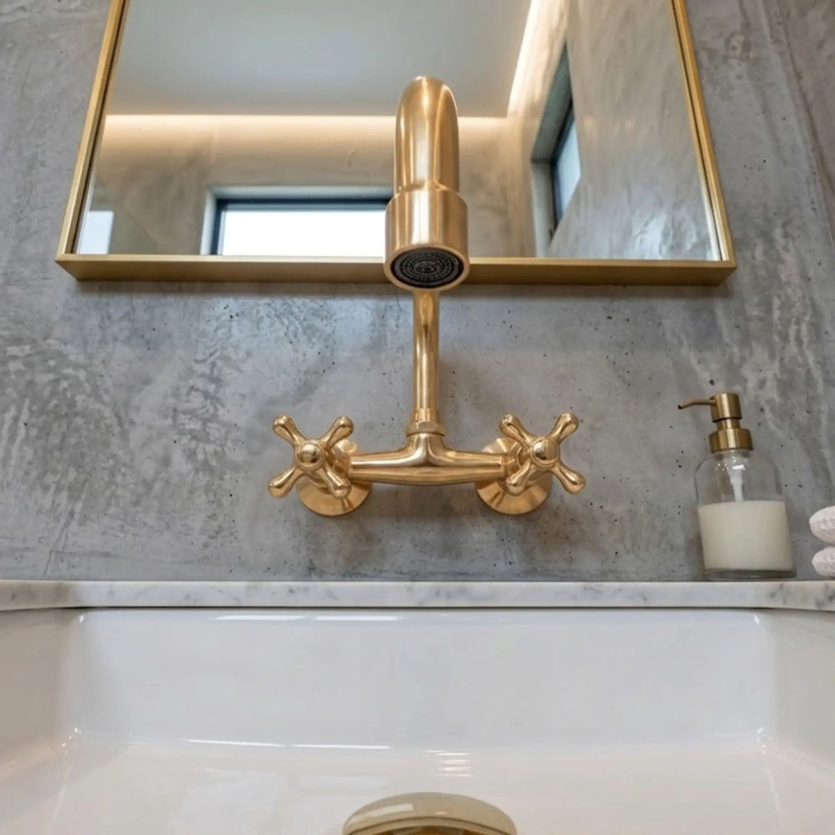Unlacquered brass wall-mount faucet with cross handles and cylindrical spout, front view on concrete wall above white sink