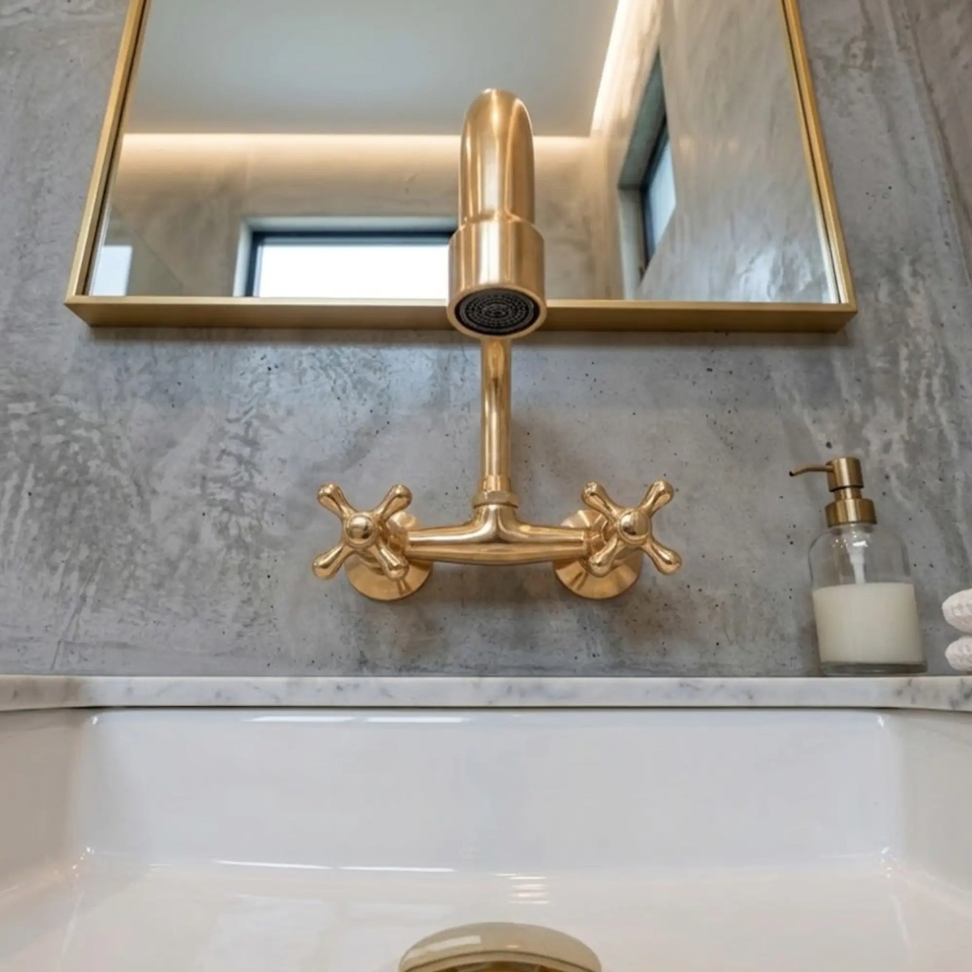 Unlacquered brass wall-mount faucet with cross handles and cylindrical spout, front view on concrete wall above white sink