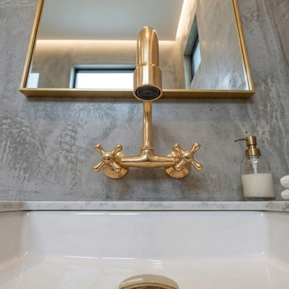 Unlacquered brass wall-mount faucet with cross handles and cylindrical spout, front view on concrete wall above white sink