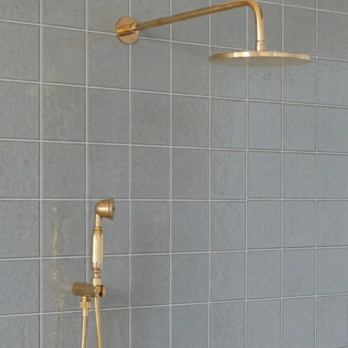 Unlacquered brass wall-mount rain shower system with handheld sprayer and exposed pipe design installed on gray tile bathroom wall