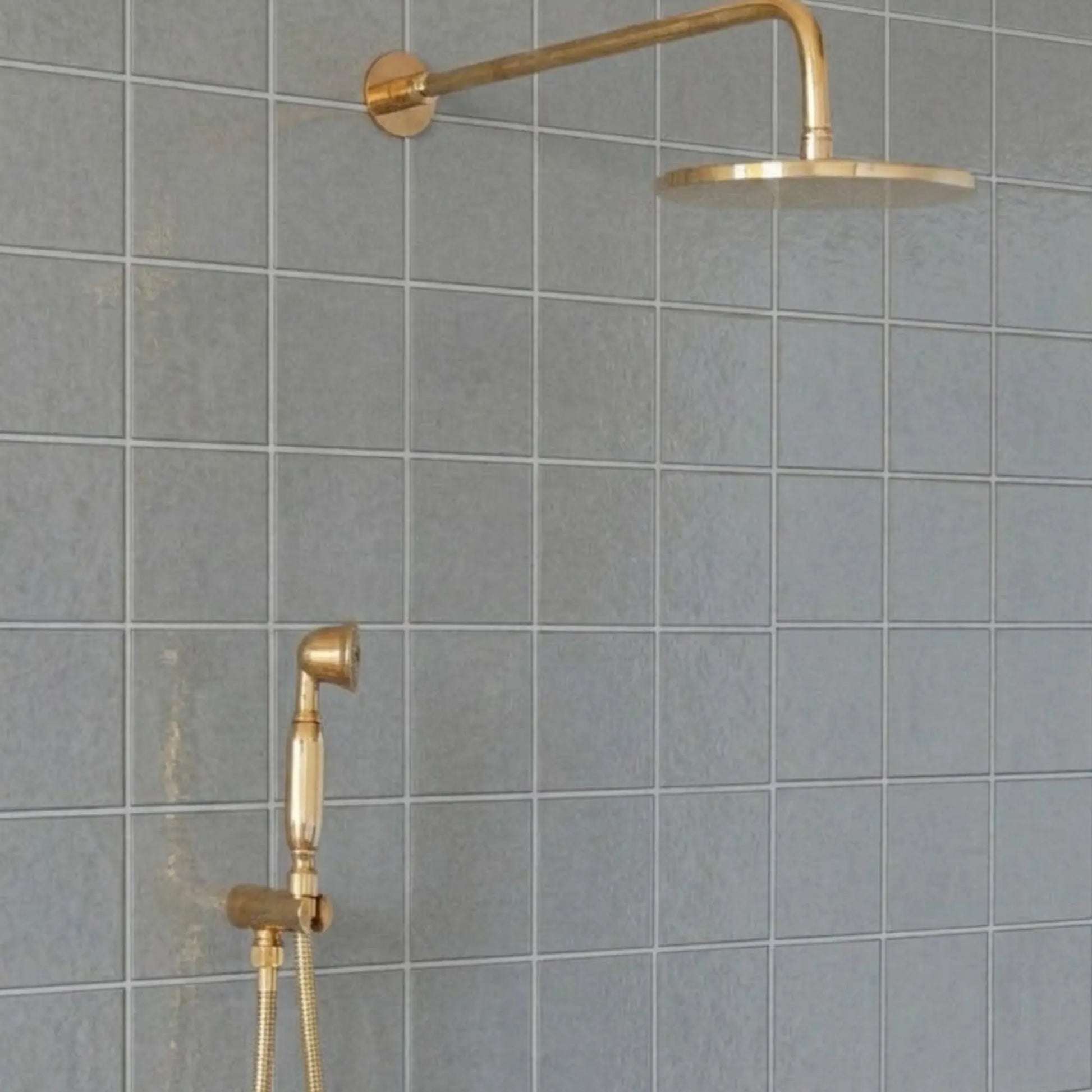 Unlacquered brass wall-mount rain shower system with handheld sprayer and exposed pipe design installed on gray tile bathroom wall