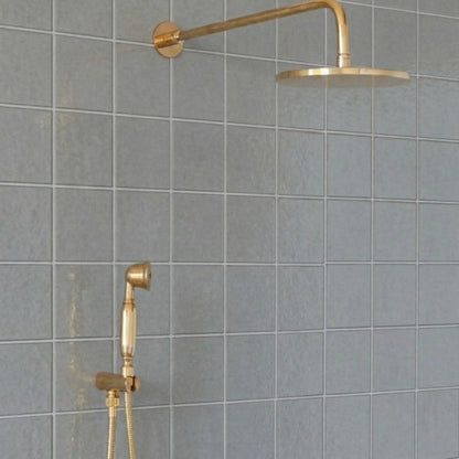 Unlacquered brass wall-mount rain shower system with handheld sprayer and exposed pipe design installed on gray tile bathroom wall