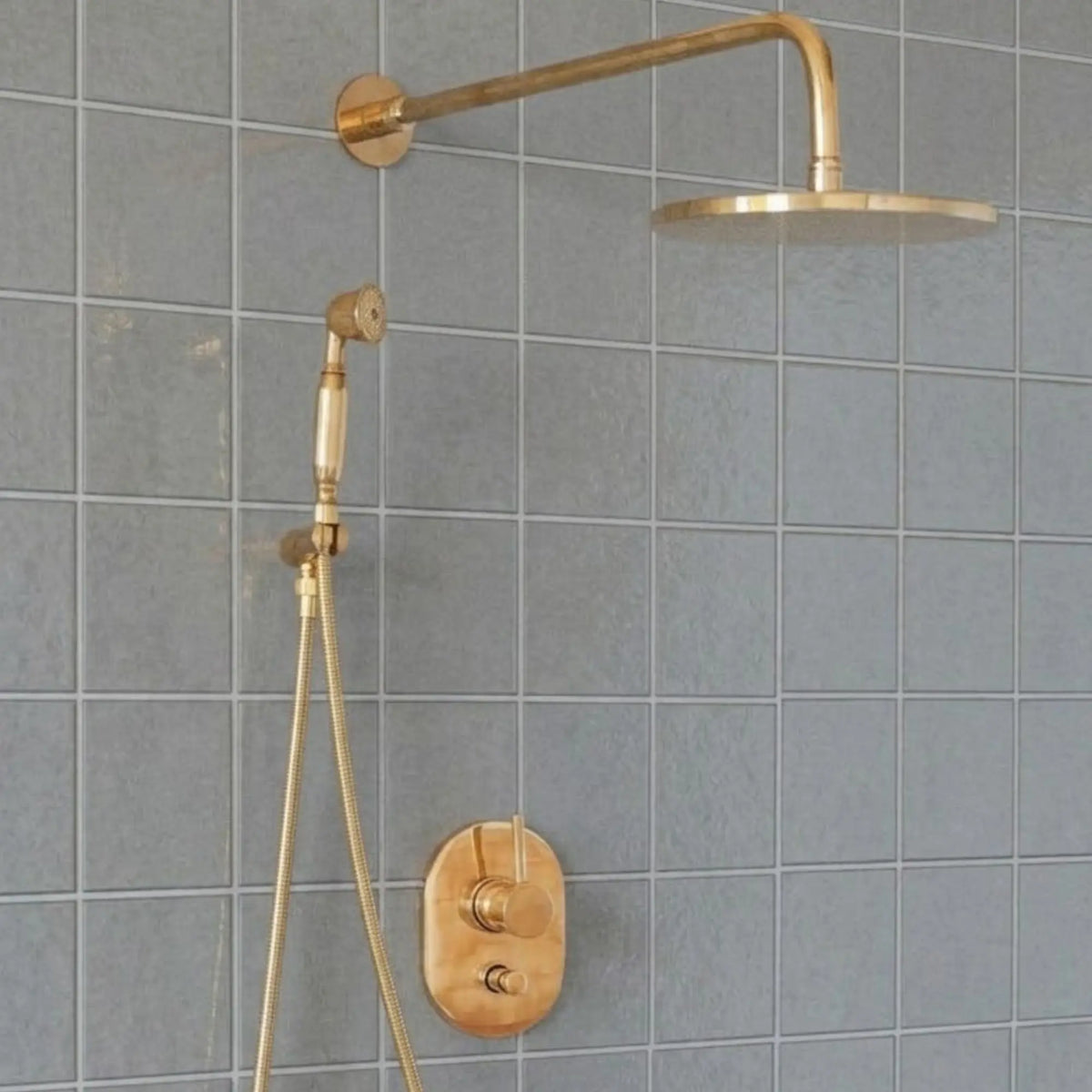 Unlacquered brass wall mount rain shower system with handheld shower and exposed pipe design installed on gray tiled bathroom wall
