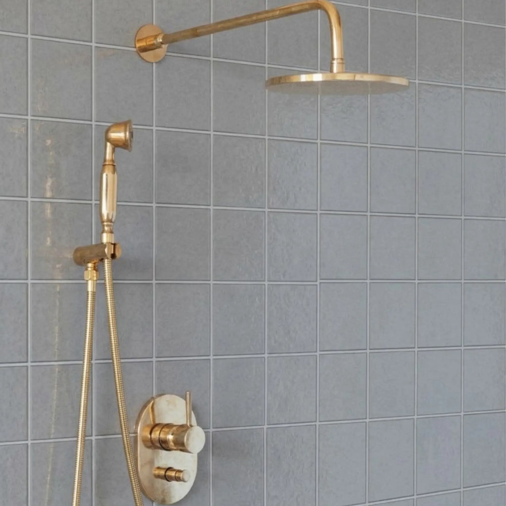 Unlacquered brass wall mount rain shower system with handheld sprayer and exposed pipe design installed on gray tiled bathroom wall