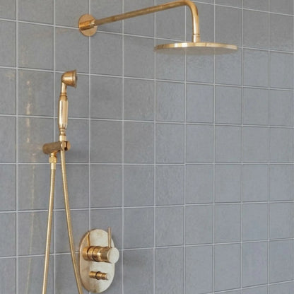 Unlacquered brass wall mount rain shower system with handheld sprayer and exposed pipe design installed on gray tiled bathroom wall