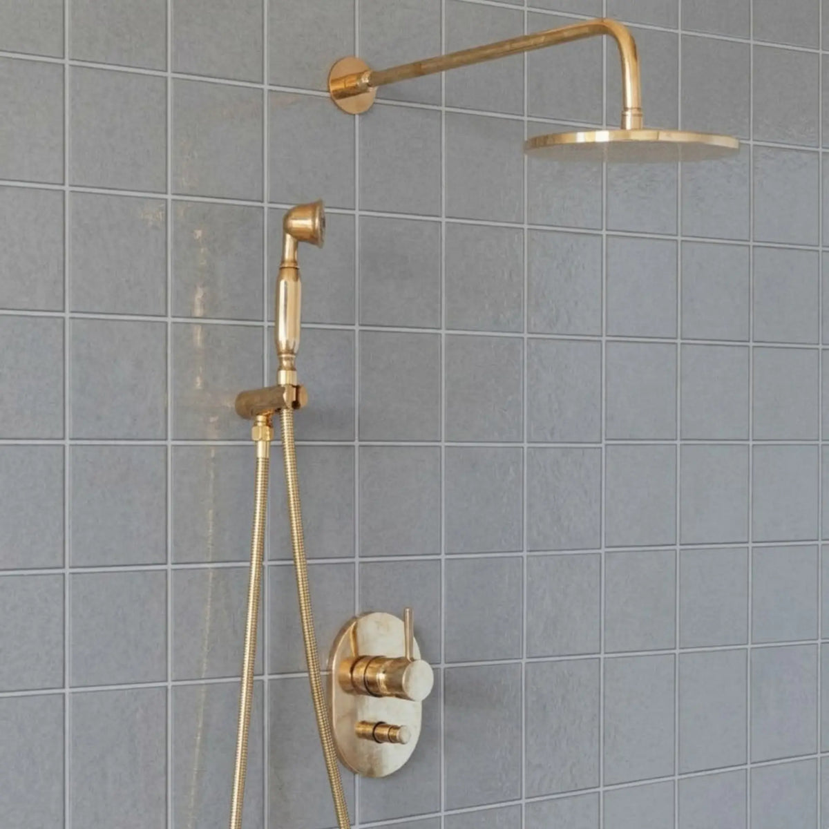 Unlacquered brass wall mount rain shower system with handheld shower and exposed pipe design installed on gray tile bathroom wall