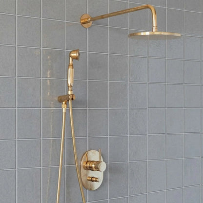 Unlacquered brass wall mount rain shower system with handheld shower and exposed pipe design installed on gray tile bathroom wall