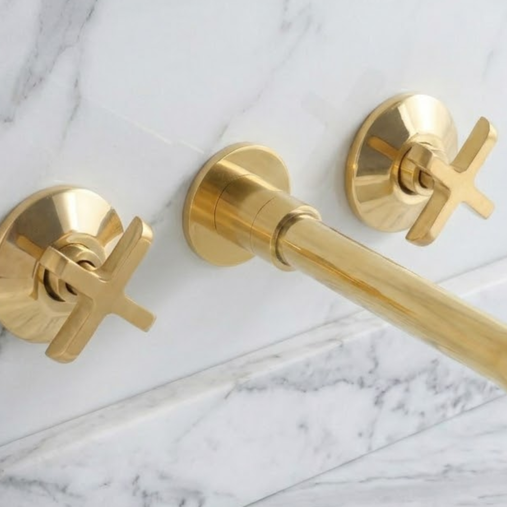 Close-up of a solid unlacquered brass wall-mounted bathroom faucet with cross handles installed on a white marble wall above a brass sink, featuring a natural living brass finish.