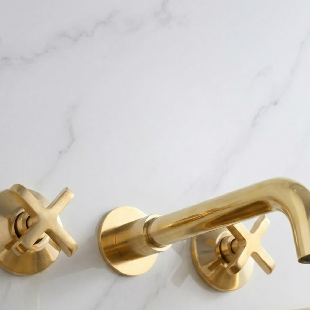 Luxury solid unlacquered brass wall-mounted bathroom faucet with cross handles installed on a white marble wall, featuring a curved spout and natural living brass finish.