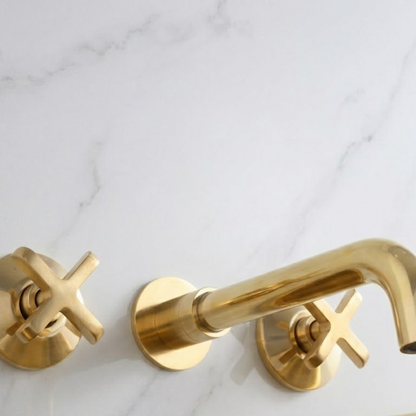 Luxury solid unlacquered brass wall-mounted bathroom faucet with cross handles installed on a white marble wall, featuring a curved spout and natural living brass finish.