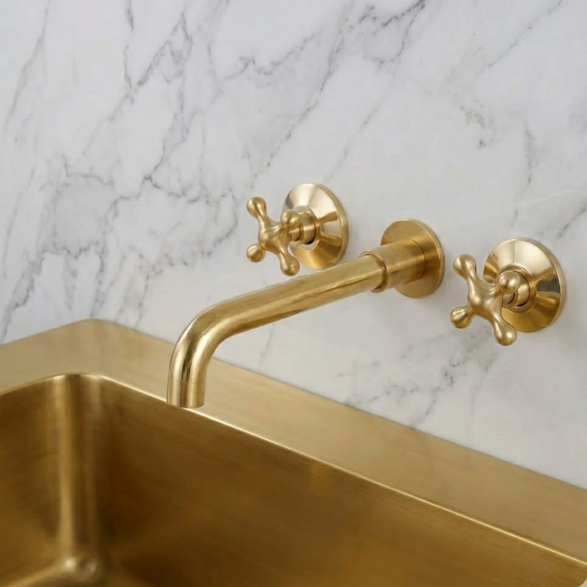 Solid unlacquered brass wall-mounted bathroom faucet with cross handles installed above a brass tub against a white marble wall, featuring a natural living patina finish.