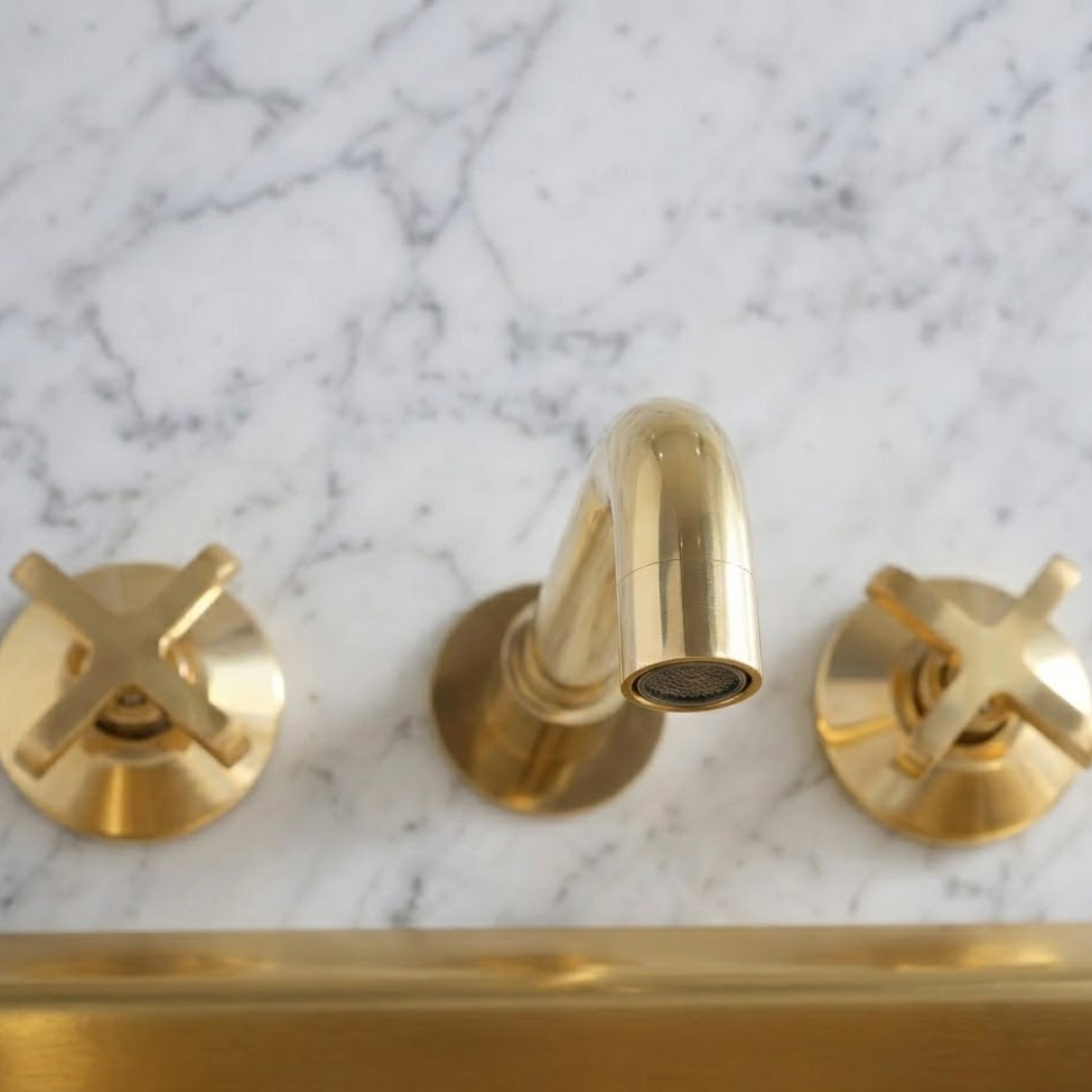 Top view of a solid unlacquered brass wall-mounted bathroom faucet with cross handles installed on a white marble wall above a brass tub, featuring a natural living finish.