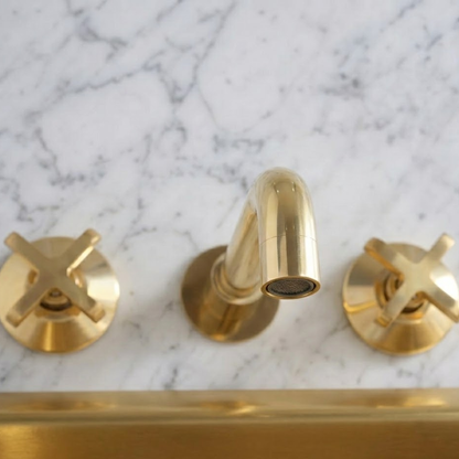 Top view of a solid unlacquered brass wall-mounted bathroom faucet with cross handles installed on a white marble wall above a brass tub, featuring a natural living finish.