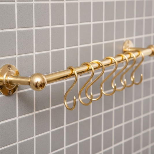 Unlacquered brass wall mounted pot rail with solid brass S hooks installed on tiled kitchen wall