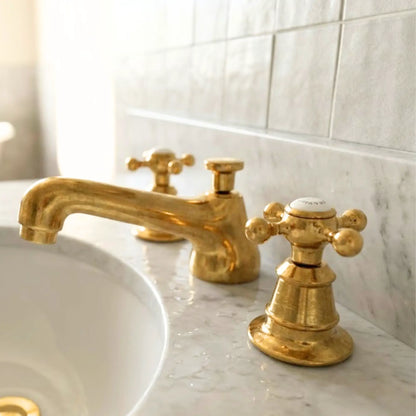 Unlacquered brass widespread bathroom faucet with cross handles on marble countertop developing natural patina