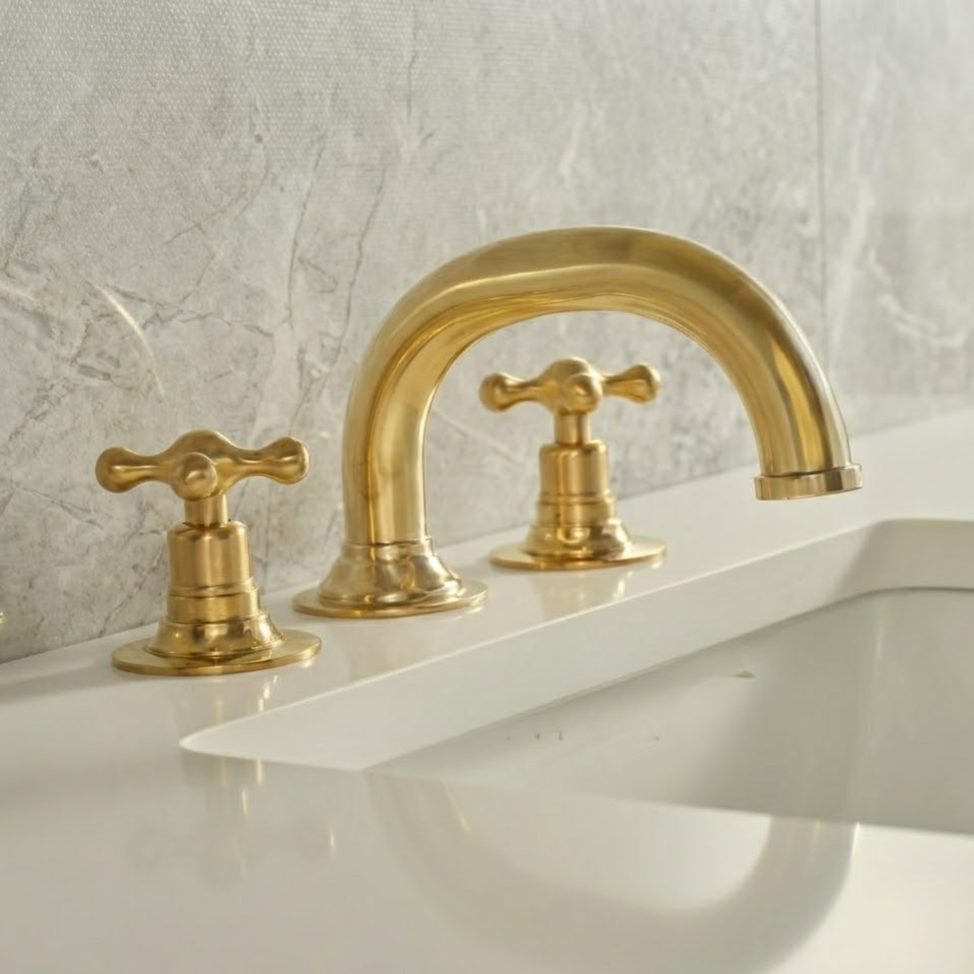 Solid unlacquered brass widespread bathroom faucet with cross handles installed on a white ceramic sink against a light gray textured wall